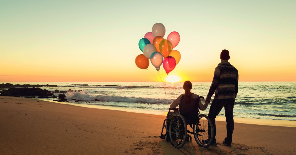 男性と風船を持った車椅子の女性の二人が日暮の海辺にいる様子。
